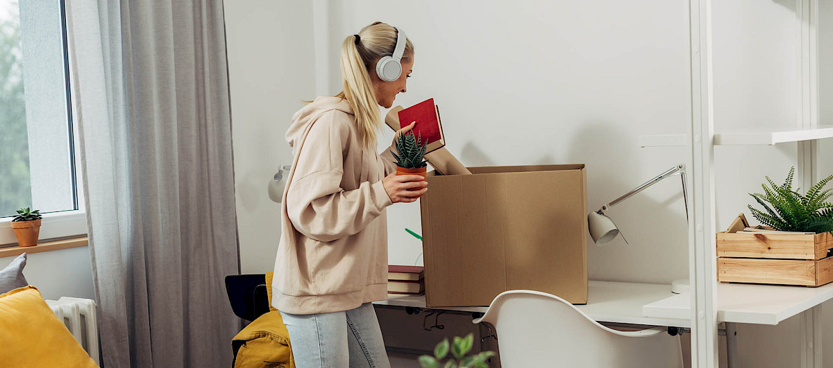 A blonde woman is packing a moving box while wearing white headphones.